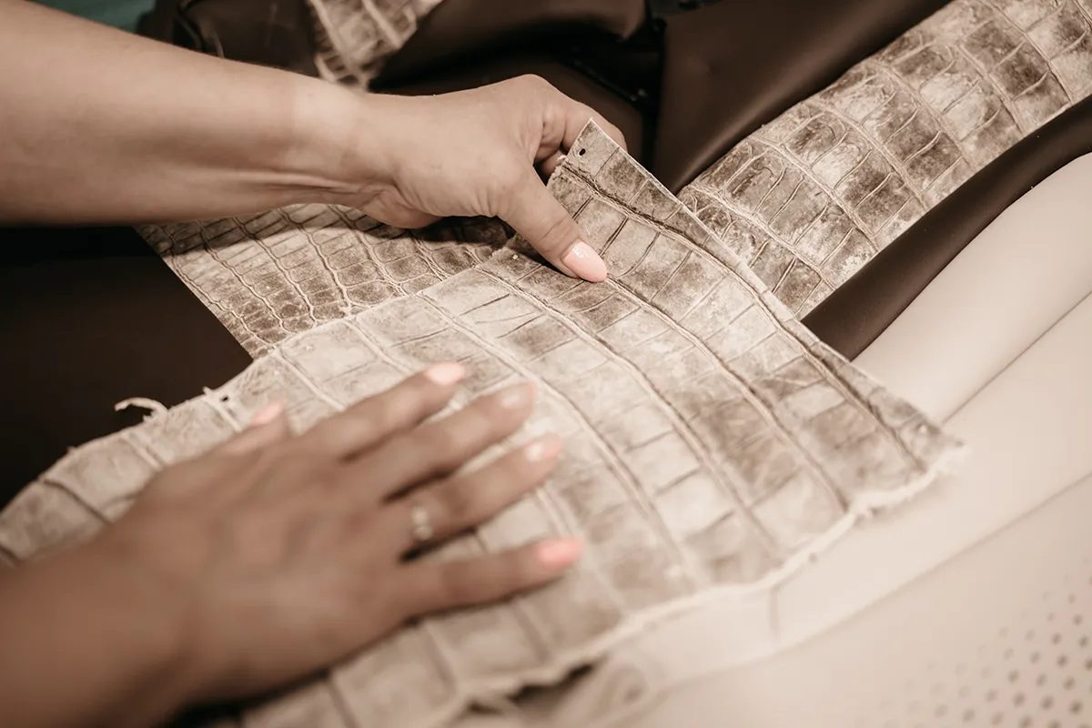 Close-up of two hands working with a piece of textured, light-colored leather, possibly alligator or crocodile skin, on top of dark brown material, suggesting craftsmanship or upholstery work.
