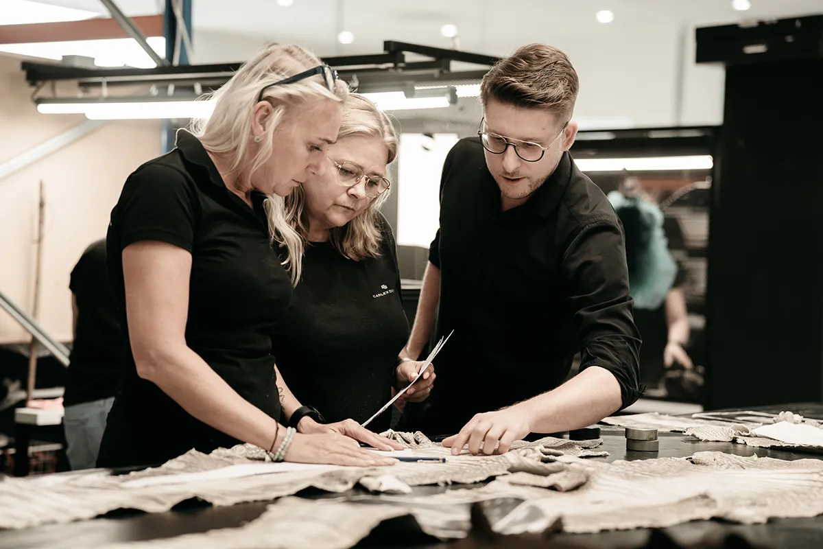 Three people stand at a table examining fabric samples. Two women and a man discuss and point to the materials; scissors and sewing tools are visible on the table, suggesting a collaborative design or tailoring process.