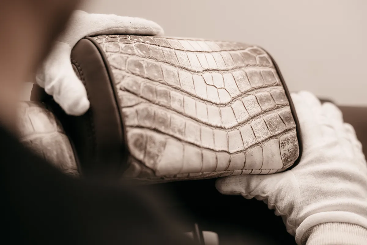 A person wearing white gloves is holding a luxury handbag with a textured crocodile leather pattern, focusing on its craftsmanship and detail.